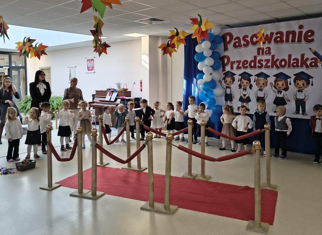 Przedszkolaki stoją w szeregu przed ścianką z napisem „Pasowanie na Przedszkolaka”. W sali przygotowano czerwony dywan, złote słupki z bordowymi sznurami oraz dekoracje z balonów i kolorowych papierowych ozdób. Dzieci ubrane są w eleganckie stroje.