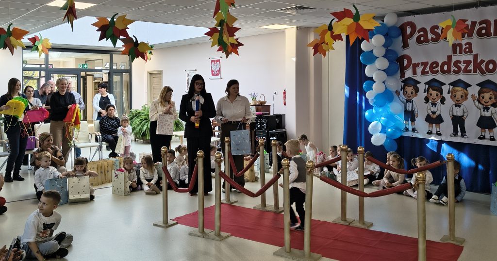 Przedszkolaki siedzą na podłodze z prezentami, a część dzieci przechodzi po czerwonym dywanie podczas uroczystości pasowania. Obok stoją przedstawicielki przedszkola, a w tle widoczna jest dekoracyjna ścianka z napisem „Pasowanie na Przedszkolaka”, niebiesko-białe balony i kolorowe ozdoby.
