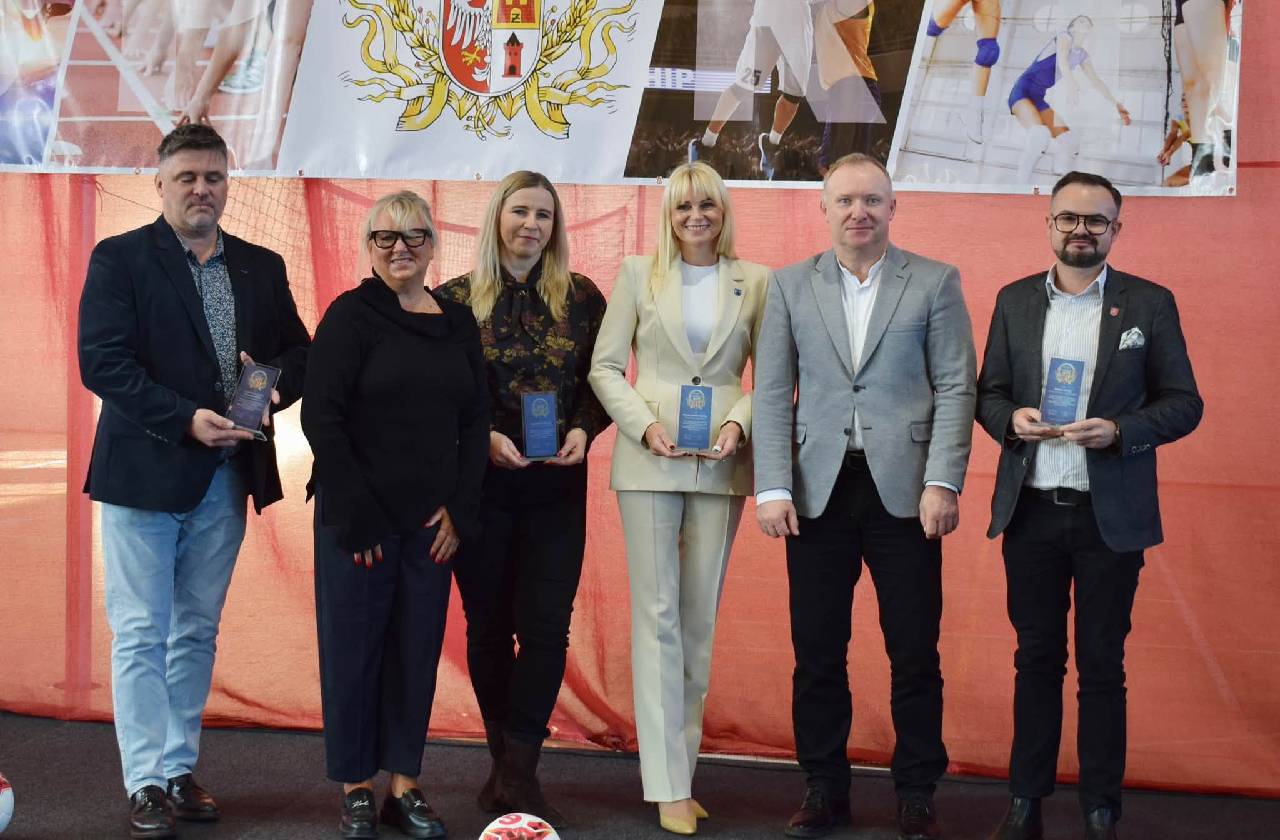 Pięć osób pozuje do wspólnego zdjęcia podczas Gali Sportu Powiatu Żyrardowskiego, prezentując pamiątkowe statuetki. Wśród uczestników znajdują się kobiety i mężczyźni ubrani w eleganckie stroje. Za nimi znajduje się baner z nazwą wydarzenia, herbem powiatu oraz fotografiami przedstawiającymi różne dyscypliny sportowe.
