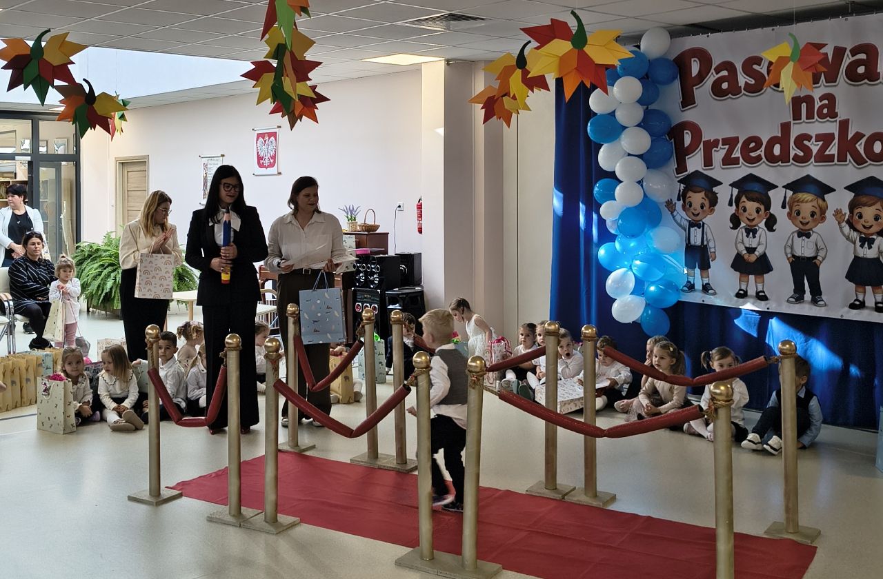 Przedszkolaki siedzą na podłodze z prezentami, a część dzieci przechodzi po czerwonym dywanie podczas uroczystości pasowania. Obok stoją przedstawicielki przedszkola, a w tle widoczna jest dekoracyjna ścianka z napisem „Pasowanie na Przedszkolaka”, niebiesko-białe balony i kolorowe ozdoby.