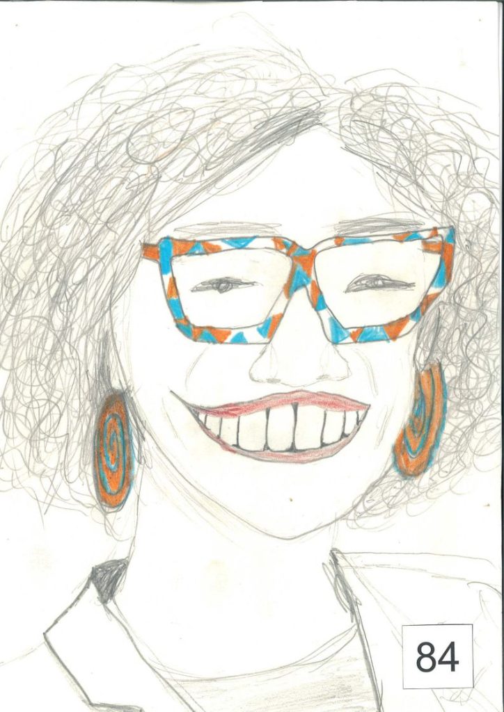 Portret karykaturalny przedstawia osobę z bujnymi, kręconymi włosami, dużymi okularami w pomarańczowo-niebieskich oprawkach oraz dużymi kolczykami w podobnych kolorach. Postać ma szeroki uśmiech z widocznymi zębami i ubrana jest w jasną koszulę lub bluzkę. Rysunek wykonano ołówkiem i kredkami na białym tle.