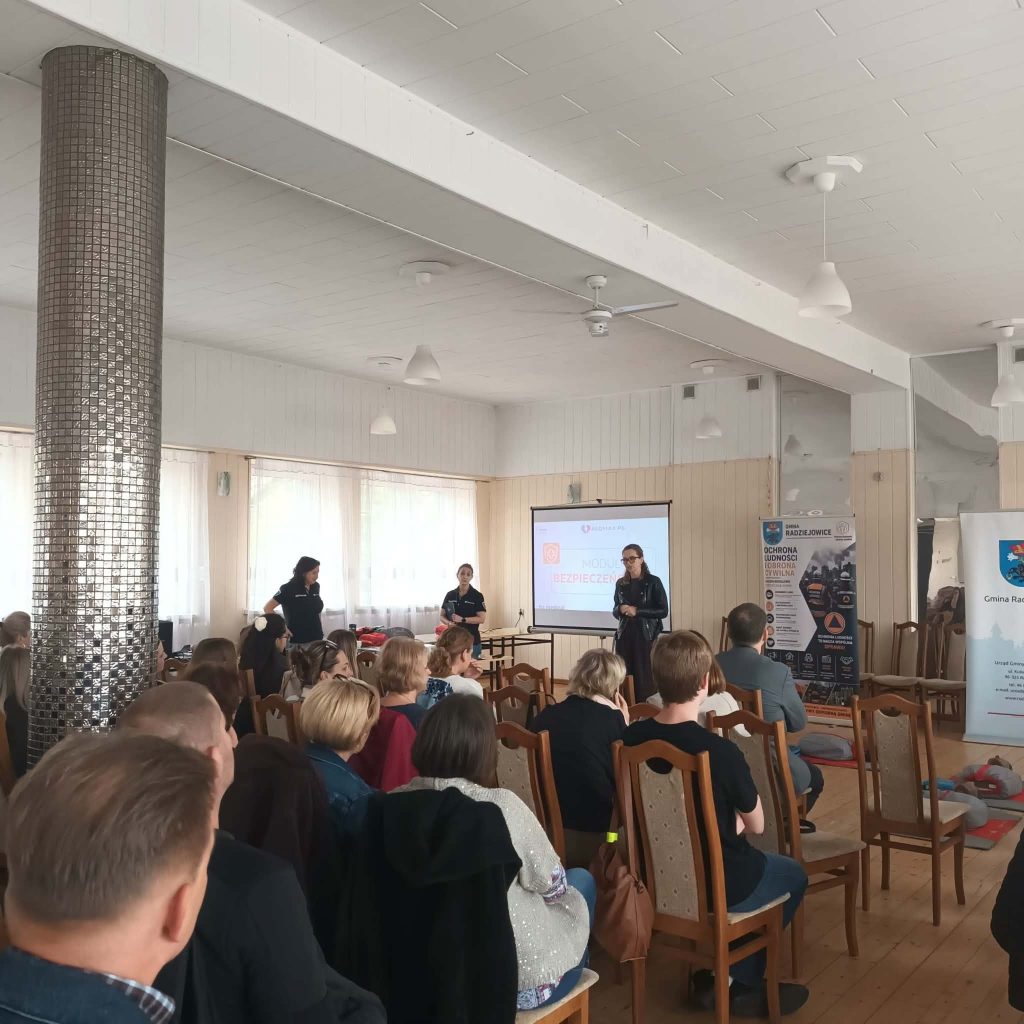 Uczestnicy szkolenia siedzą na krzesłach i słuchają prowadzących. Na ekranie wyświetlana jest prezentacja, a obok znajdują się roll-upy informacyjne dotyczące ochrony ludności i obrony cywilnej Gminy Radziejowice.