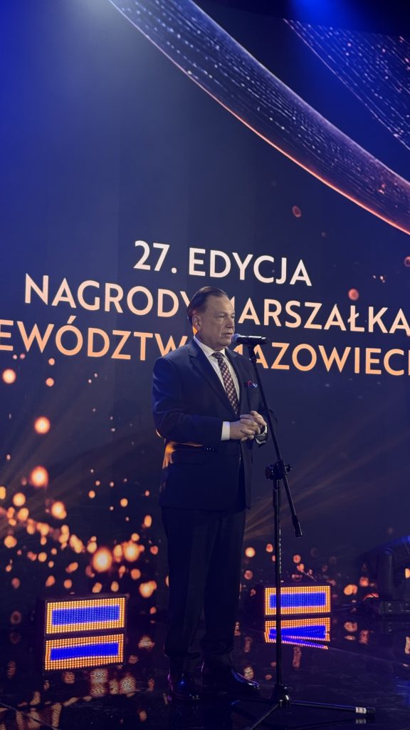 Mężczyzna w granatowym garniturze i krawacie przemawia przy mikrofonie na scenie podczas 27. edycji Nagrody Marszałka Województwa Mazowieckiego. W tle widoczny jest ekran z nazwą wydarzenia i dekoracyjną oprawą świetlną w odcieniach granatu oraz pomarańczu.