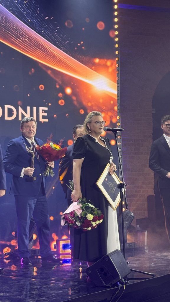 Kobieta w czarnej sukni z białymi wstawkami odbiera dyplom i bukiet kwiatów podczas uroczystości wręczenia Nagrody Marszałka Województwa Mazowieckiego. Za nią stoją mężczyźni w garniturach, w tym laureat trzymający statuetkę i bukiet kwiatów. Uroczystość odbywa się na oświetlonej scenie.