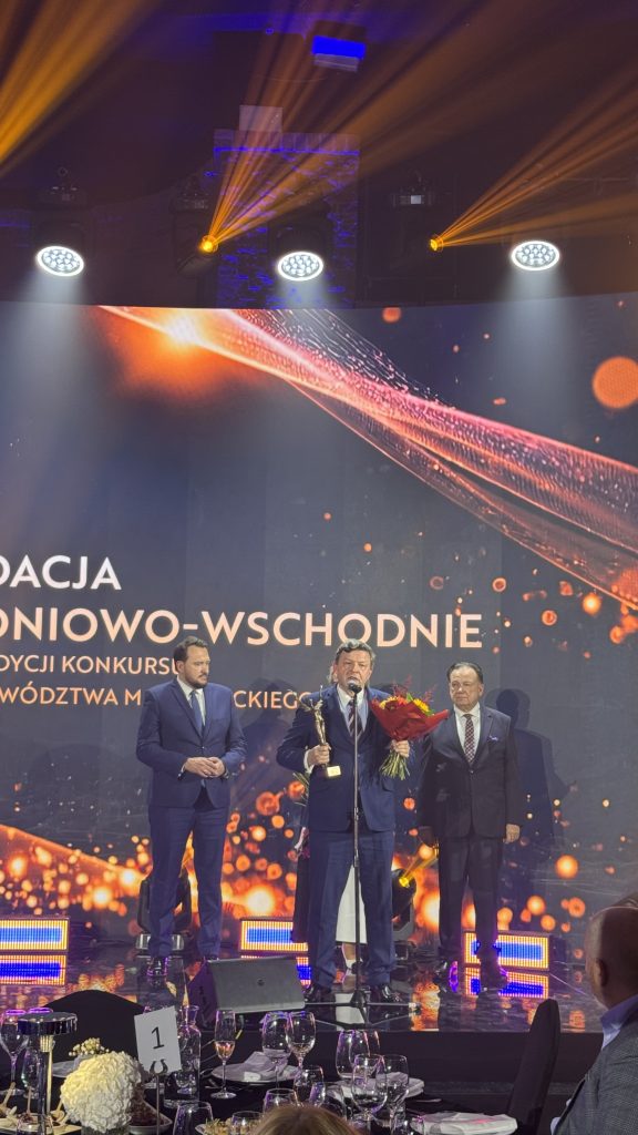 Trzech mężczyzn w garniturach stoi na scenie podczas gali wręczenia Nagrody Marszałka Województwa Mazowieckiego. Mężczyzna pośrodku przemawia do mikrofonu, trzymając statuetkę i bukiet kwiatów. W tle widoczny jest ekran z nazwą wydarzenia i pomarańczową oprawą graficzną.