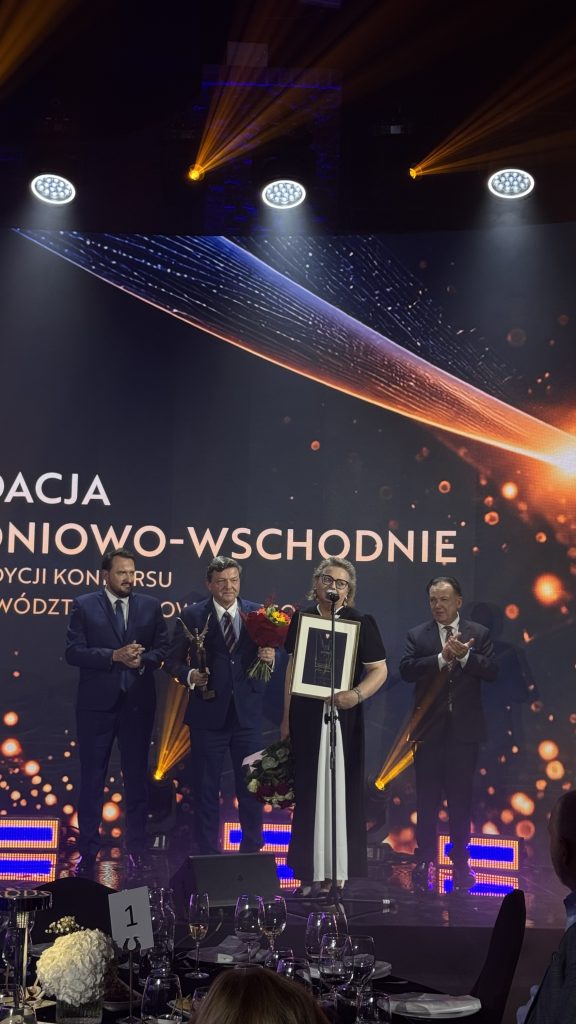 Kobieta w czarno-białej sukni, trzymająca oprawiony dyplom i bukiet kwiatów, stoi przy mikrofonie na scenie w towarzystwie mężczyzn w garniturach. Pozostali uczestnicy uroczystości gratulują laureatom. W tle widoczna jest dekoracyjna oprawa świetlna gali.