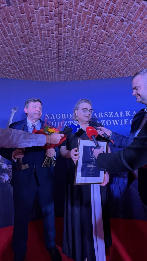 Kobieta i mężczyzna, laureaci Nagrody Marszałka Województwa Mazowieckiego, udzielają wywiadu przedstawicielom mediów. Kobieta trzyma oprawiony dyplom, a mężczyzna statuetkę i bukiet kwiatów. Dziennikarze kierują w ich stronę mikrofony. W tle znajduje się ściana z nazwą nagrody.