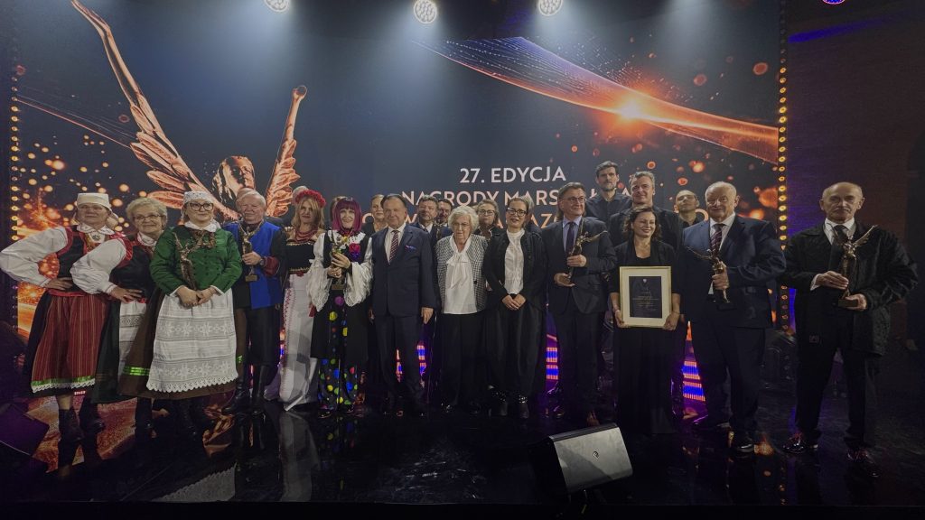 Laureaci i uczestnicy 27. edycji Nagrody Marszałka Województwa Mazowieckiego pozują do wspólnego zdjęcia na scenie. W grupie znajdują się osoby w strojach regionalnych oraz eleganckich strojach wizytowych. Część uczestników trzyma statuetki, a jedna z osób oprawiony dyplom. W tle widoczny jest ekran z nazwą wydarzenia.
