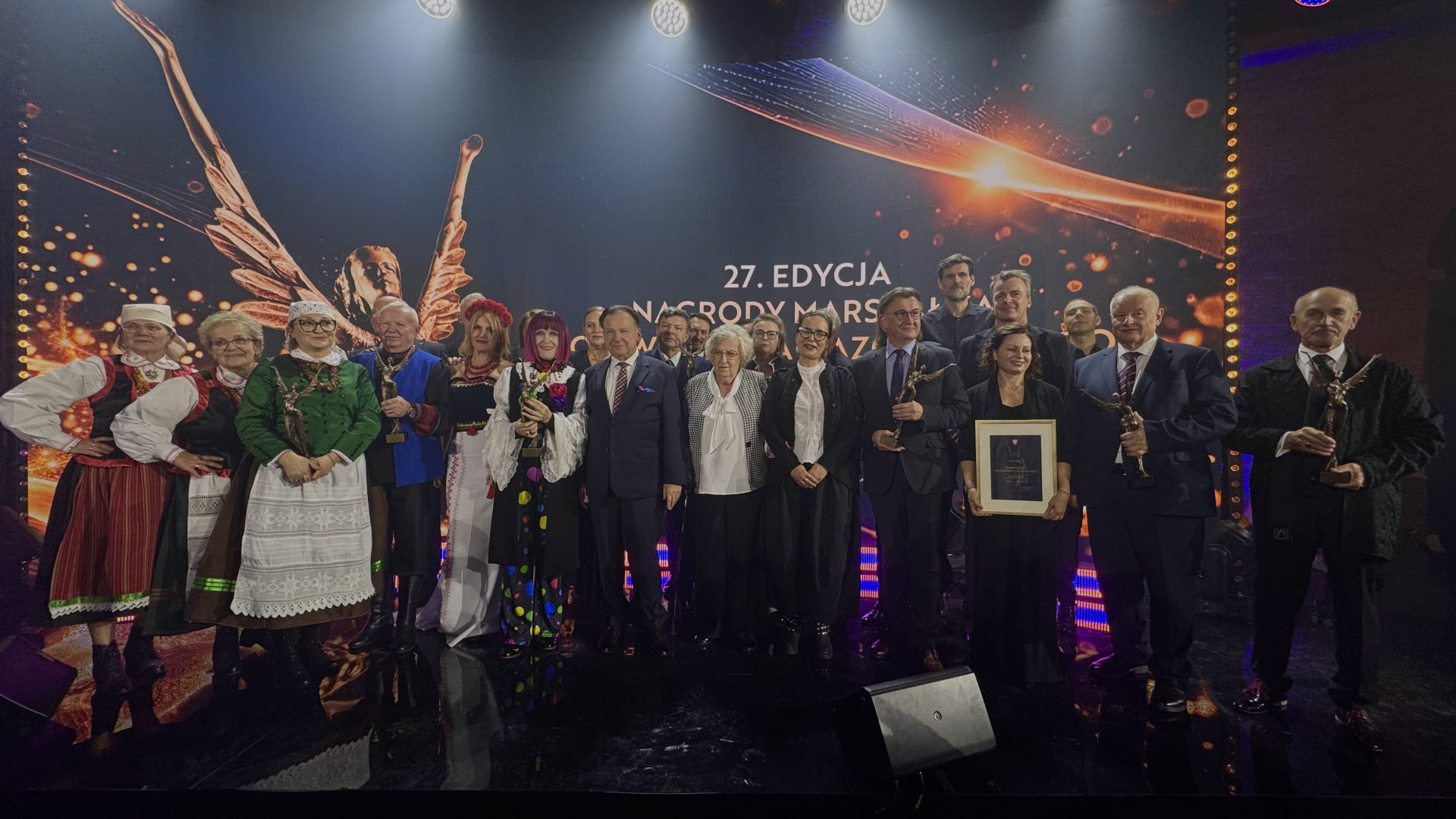 Laureaci i uczestnicy 27. edycji Nagrody Marszałka Województwa Mazowieckiego pozują do wspólnego zdjęcia na scenie. W grupie znajdują się osoby w strojach regionalnych oraz eleganckich strojach wizytowych. Część uczestników trzyma statuetki, a jedna z osób oprawiony dyplom. W tle widoczny jest ekran z nazwą wydarzenia.